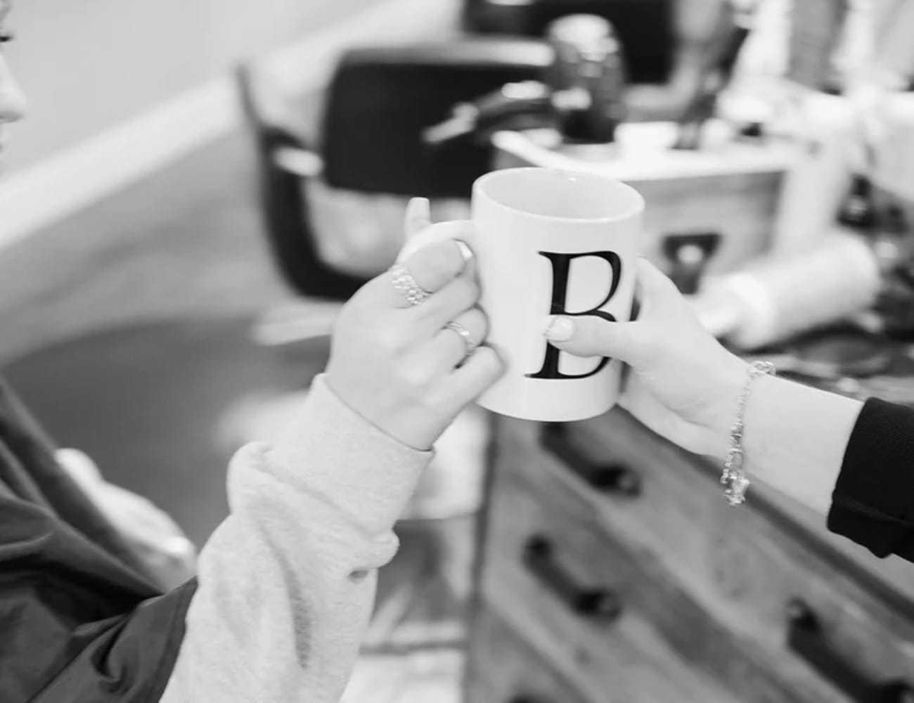 Two people clink mugs, one with a letter 'B'. Cozy indoor setting.